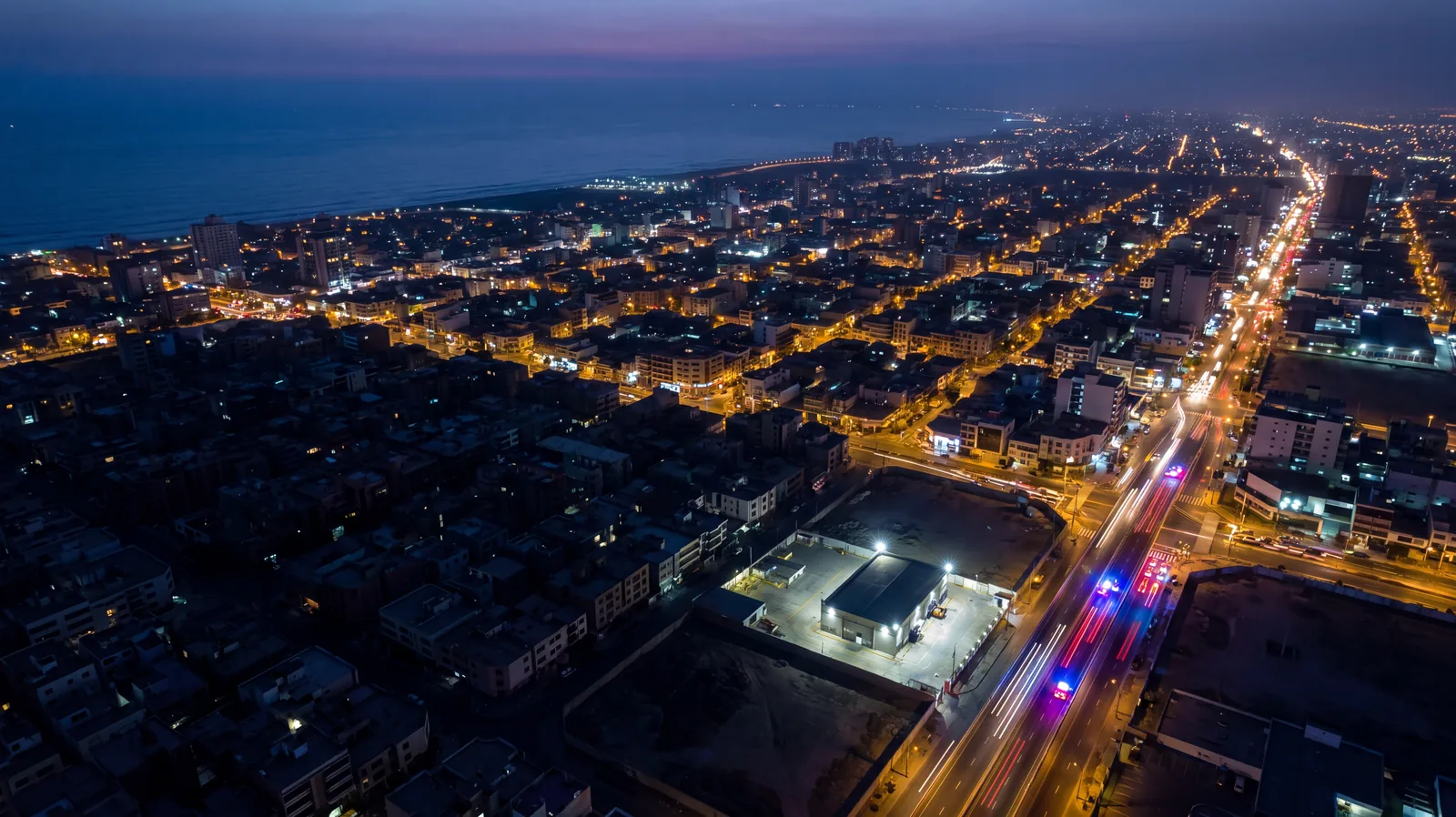 Aerial view of a coastal city at dusk where one district has lost power, with emergency vehicle light trails crossing a bridge toward it